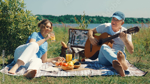 A middle-aged couple, a man and a woman, play the guitar and talk cheerfully at a picnic on a sunny summer day on the river bank.
