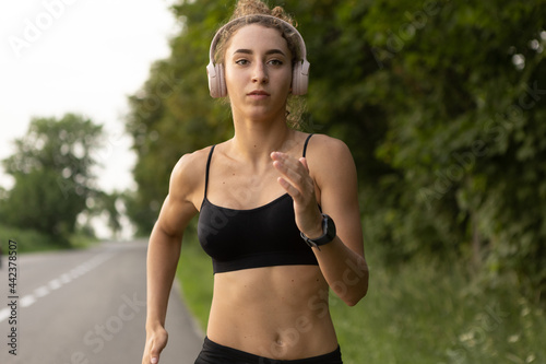Young female runner, athlete is jogging at road in summer sunshine. Beautiful caucasian woman training, listening to music. Concept of sport, healthy lifestyle, movement, activity.