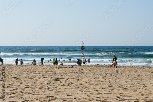 Fotografie A beach filled with beach goers around a designated swimming area sign on a sunn