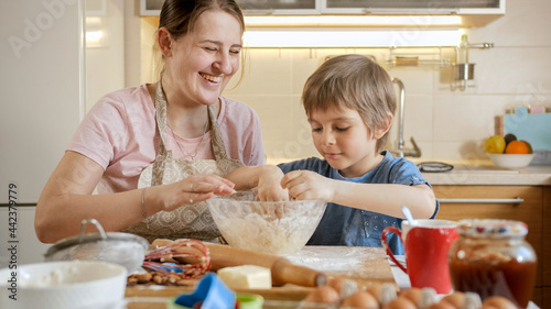 Happy smiling mother with little son kneading and smelling dough in glass bowl. Children cooking with parents, little chef, family having time together, domestic kitchen.
