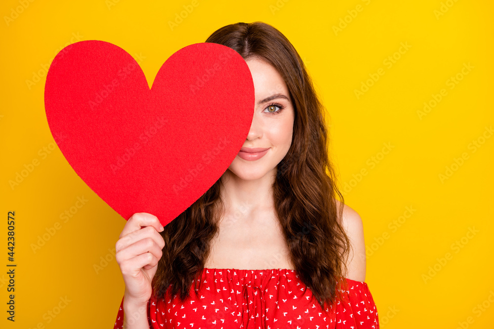 Portrait of charming young girl arm hold heart symbol card covering one eye isolated on yellow color background Portrait of charming young girl arm hold heart symbol card covering one eye isolated on yellow color background