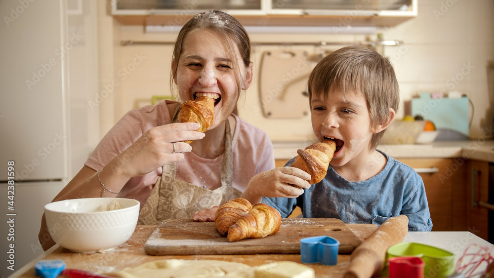 Happy smiling boy with mother covered in flour eating tasty croissant after baking at home. Children cooking with parents, little chef, family having time together, domestic kitchen. Happy smiling boy with mother covered in flour eating tasty croissant after baking at home. Children cooking with parents, little chef, family having time together, domestic kitchen.