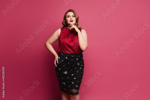 Young plus size caucasian model woman in red satin blouse and skirt posing over pink background. Gorgeous plump woman in modish outfit over pink background