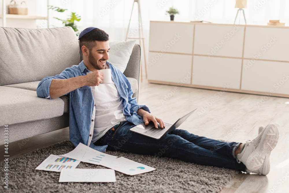 Smiling jewish man watching video on pc, drinking hot coffee