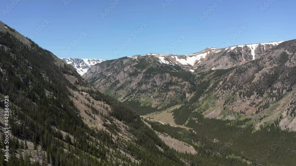 Flying above high steep slopes into the valley in high mountains with peaks partial covered with snow, clear blue sky, Montana Flying above high steep slopes into the valley in high mountains with peaks partial covered with snow, clear blue sky, Montana