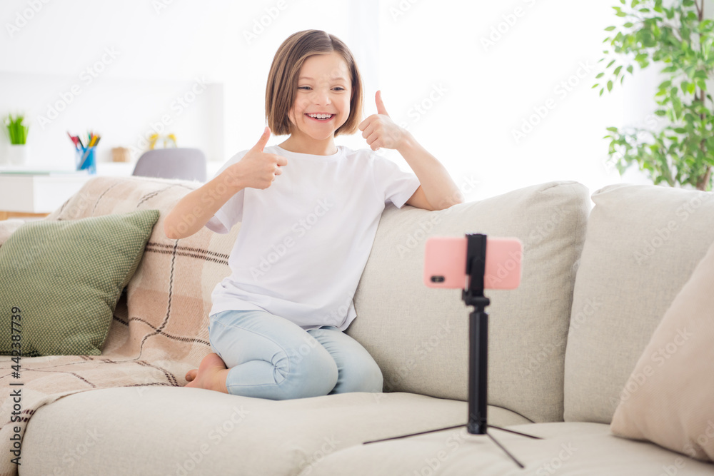 Full body photo of young little girl happy positive smile speak video call cellphone show thumbs-up cool fine choice sit couch indoors Full body photo of young little girl happy positive smile speak video call cellphone show thumbs-up cool fine choice sit couch indoors