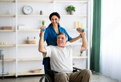 Post-stroke rehabilitation. Female physiotherapist helping elderly male patient in wheelchair to make exercises at home