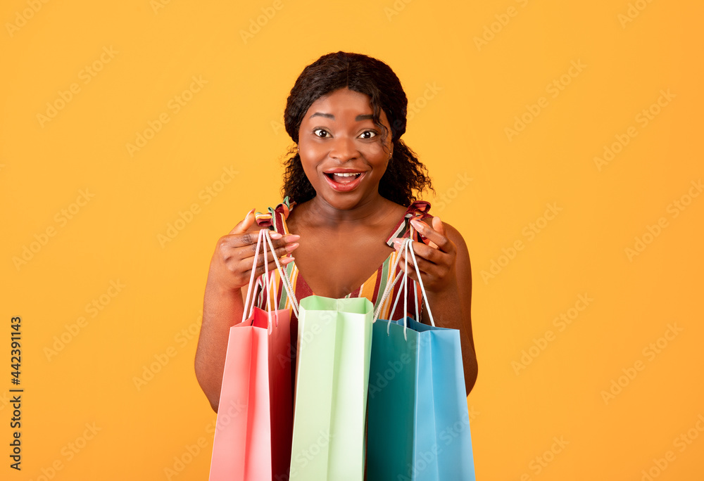 Summer shopping concept. Surprised black woman with paper bags looking at camera on orange studio background Summer shopping concept. Surprised black woman with paper bags looking at camera on orange studio background