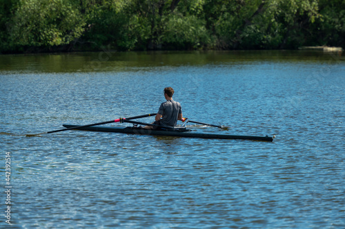 Scullers sculling to propel their long narrow boats through the water on a lake surrounded by thick vegetation and green trees. Some crews are using two oars and other individuals are use one oar