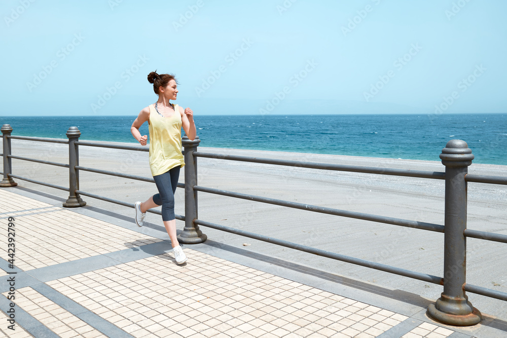 Slim Woman running on the promenade with the beach and blue sea in background. Strengthening muscles of legs and coordination of body. Practicing sport outdoors. Slim Woman running on the promenade with the beach and blue sea in background. Strengthening muscles of legs and coordination of body. Practicing sport outdoors.