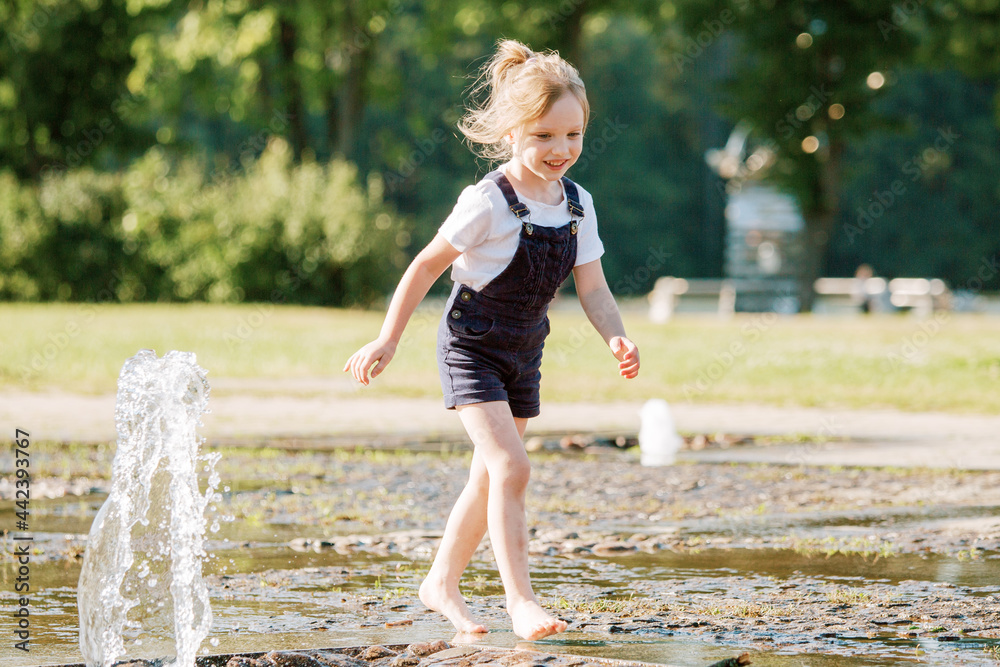 Cute cheerful girl playing in fountain. Kid in denim overall having fun in summer park