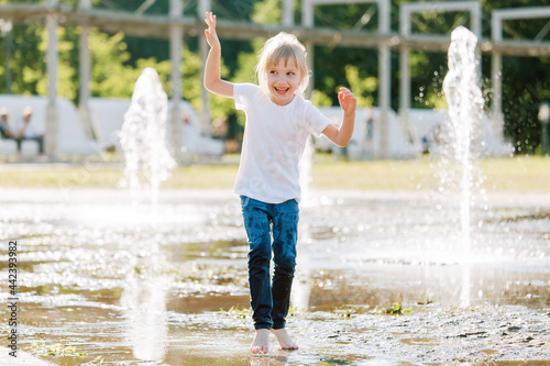 Cute cheerful girl playing in fountain. Kid in white shirt having fun in summer park