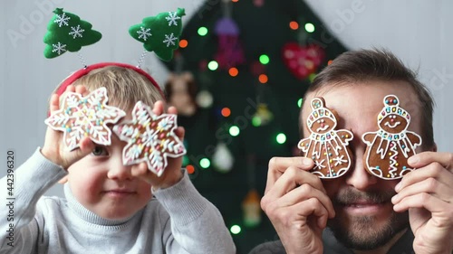 Cheerful father and son having fun near alternative christmas tree holding gingerbread cookies near eyes.