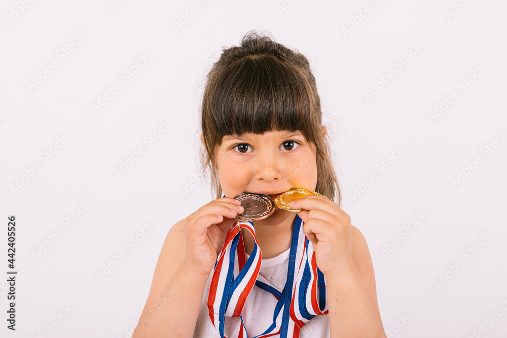 Little brown-haired girl with sports champion medals, biting them. Sport and victory concept Little brown-haired girl with sports champion medals, biting them. Sport and victory concept
