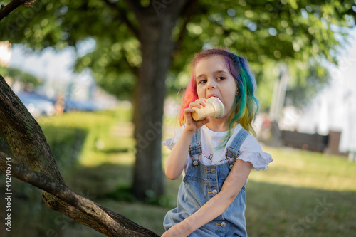 Happy little girl with colorful dyed hair eating ice cream outdoors in park. Image with selective focus