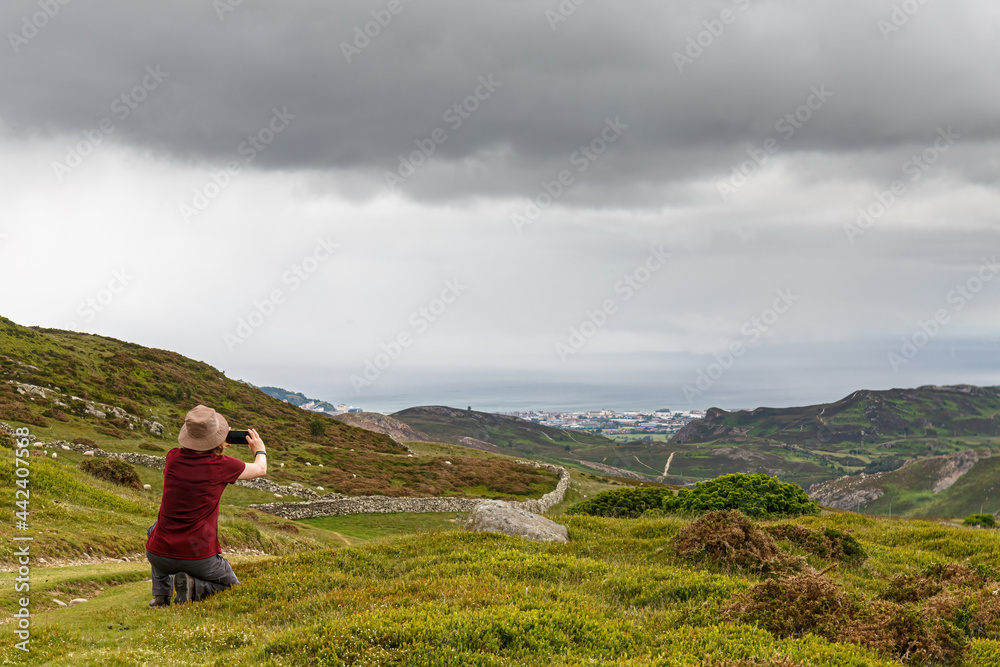 A single female walker taking photographs using a mobile phone on the scenery on the Wales Coastal Path at Penmaenmawr, Cony, Wales