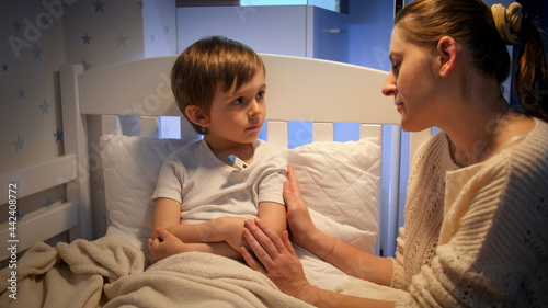 Little sick boy measuring body temperature at night and looking at young mother sitting next to him. Concept of children illness, disease and parent care