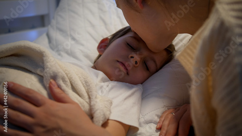 Cute little boy sleeping while caring mother kissing him and adjusting blanket. Concept of parenting and family happiness.
