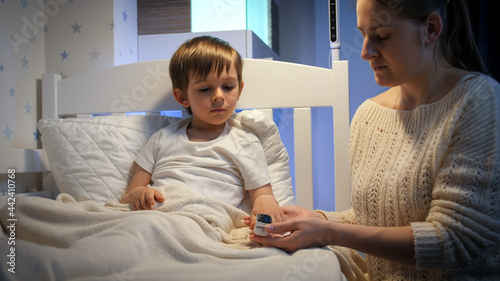 Caring mother checking level of blood oxygen of her sick little son lying in bed. Concept of child virus, domestic treatment and kids protection during coronavirus Covid-19 pandemic.