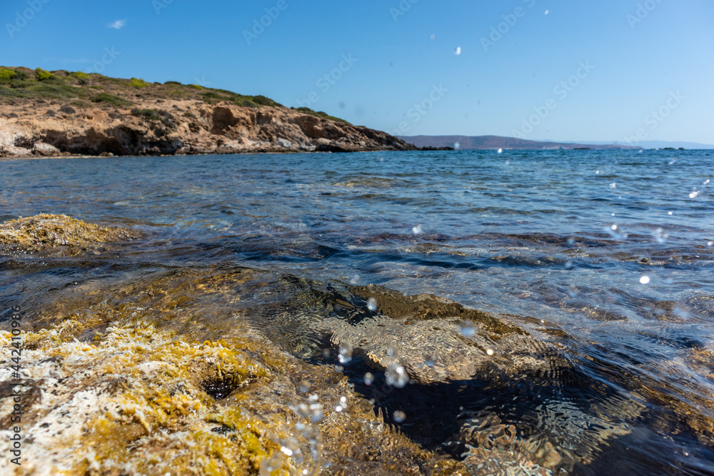 Water splash on wild mediterranean sea rocky coast with seaweed, distant cliffs and blue clear water. Travel Greece near Athens. Summer nature scenic lagoon