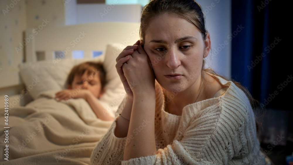Upset and stressed mother crying next to bed of her little son sleeping at night. Concept of lonely mother, family problems and maternal depression - obrazy, fototapety, plakaty 