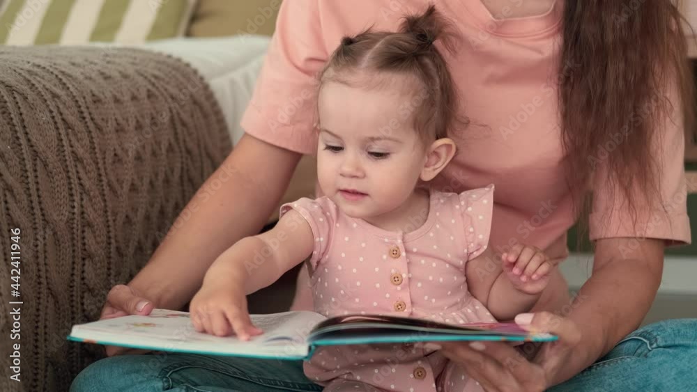 Childcare at Home, Child Protection, New Life, Leisure with Baby. Mom reads a book to the kid while sitting on the floor in the bedroom Childcare at Home, Child Protection, New Life, Leisure with Baby. Mom reads a book to the kid while sitting on the floor in the bedroom