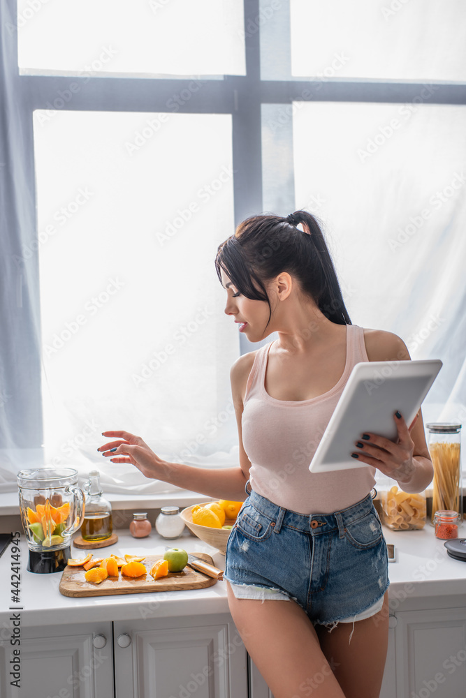 brunette young woman holding digital tablet and looking at fruits in kitchen