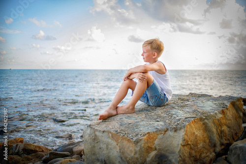 A boy in a white T-shirt and denim shorts sits on a rock near the seashore