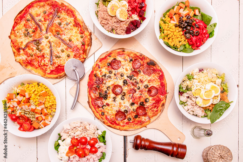 Delicious pepperoni and seafood pizzas and bowl with various types of healthy salads on white wooden table