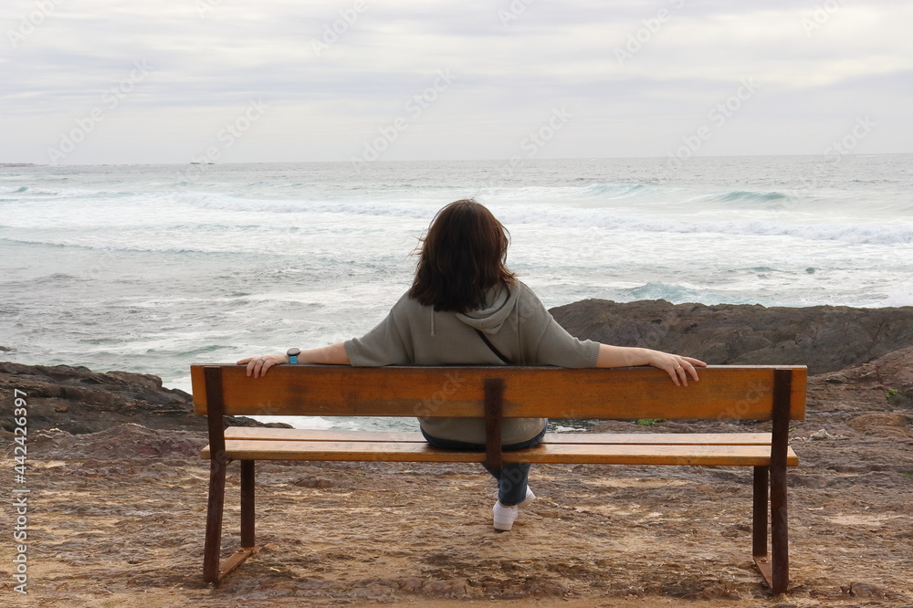 Mujer sola sentada en un banco de madera, mirando al mar en la playa de Castos de Baroña, Galica.