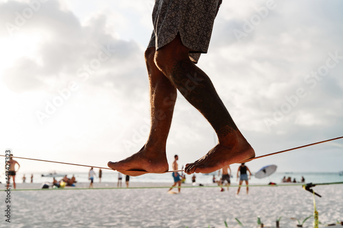 legs of a stranger performing slackline on a tight, tensioned tightrope between two palm trees in the middle of the beach overlooking the sea, sport concept