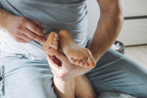 Children's feet in hands of father. Dad tickles feet of kid sitting on them. Family, having fun at home