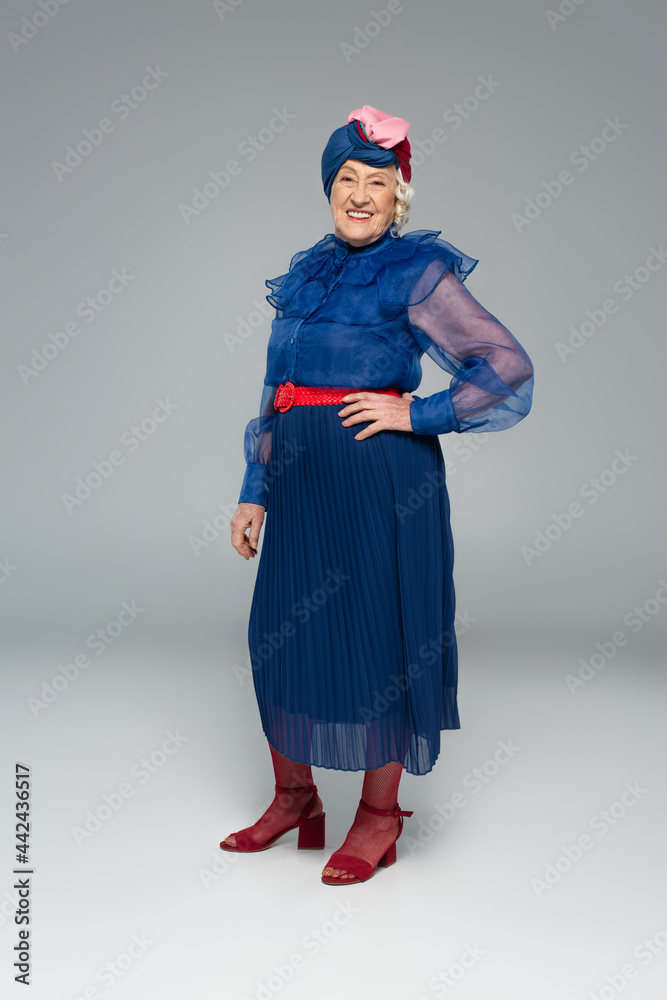 smiling elderly woman in blue dress and turban posing with hand on hip on grey smiling elderly woman in blue dress and turban posing with hand on hip on grey