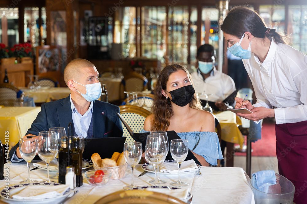 Waitress in face mask receiving order from couple of guests in masks choosing drinks and meals in restaurant