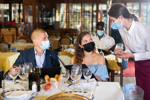 Waitress in face mask receiving order from couple of guests in masks choosing drinks and meals in restaurant