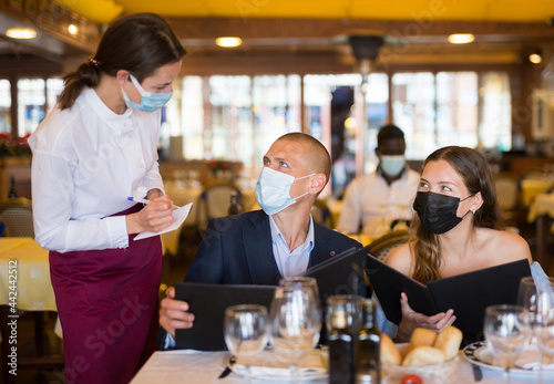 Woman waiter in protective mask is taking order from clients in restaurante indoor