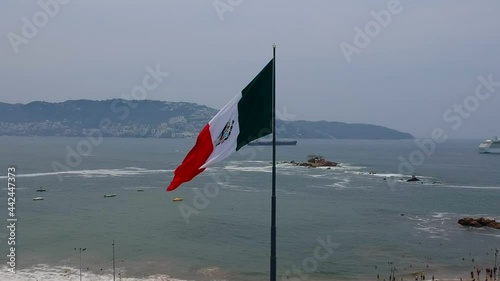 Acercamiento aéreo a bandera de México ondeando con el viento con el mar y rocas de fondo photo