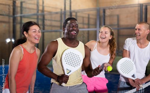 Multiethnic group of cheerful paddle tennis players standing on closed court after match, friendly talking. © JackF