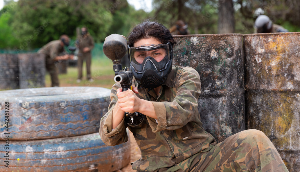 Fototapeta premium Female paintball player in camouflage and mask aiming with gun in shootout outdoors
