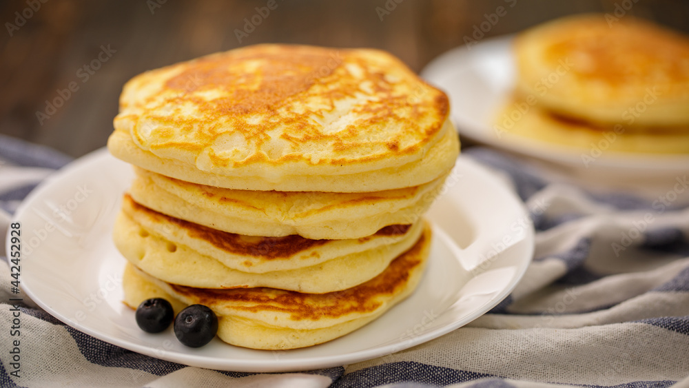 Stack of delicious pancakes sweet food with blueberries and blur dry leaves, plaid fabric in white plate on wooden background.