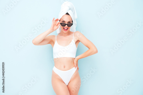 Young beautiful smiling woman in white lingerie. Sexy carefree model in underwear and towel on head posing near light blue wall in studio. Positive and happy female enjoying morning in sunglasses