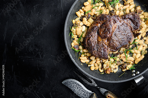 Rib eye steak with herbs, roasted beans, thyme and garlic, on frying cast iron pan, on black stone background, top view flat lay, with copy space for text