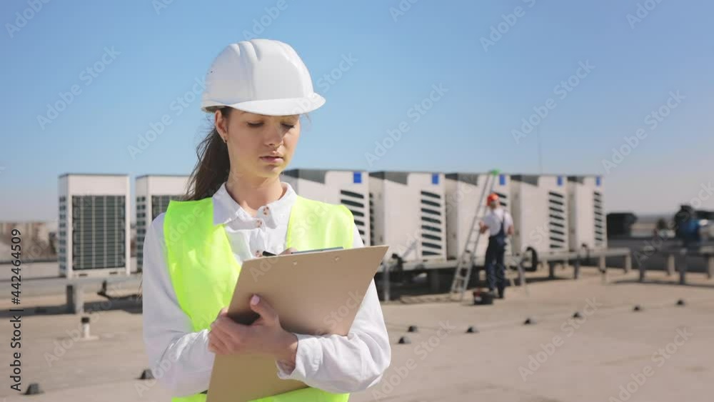 Portrait of a young woman engineer who is making entries in documents and watching the engineer's work. The engineer is taking tools to fix the air conditioners in the background. They are wearing Portrait of a young woman engineer who is making entries in documents and watching the engineer's work. The engineer is taking tools to fix the air conditioners in the background. They are wearing