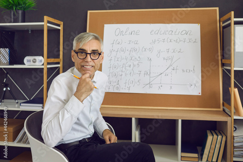 Portrait of a senior positive online math teacher sitting at a board with math formulas. Caucasian male teacher who teaches students remotely. Teaching, online education and lifestyle concept.