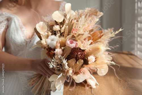 Bride holding beautiful dried flower bouquet near window at home, closeup
