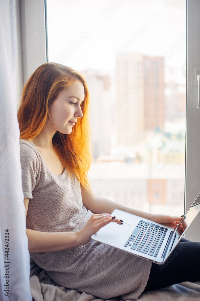 Young attractive brunette sitting on the windowsill, with laptop on her lap, typing on keyboard. Beautiful long-haired woman works at home, in city apartment. Vertical photo. Young attractive brunette sitting on the windowsill, with laptop on her lap, typing on keyboard. Beautiful long-haired woman works at home, in city apartment. Vertical photo.