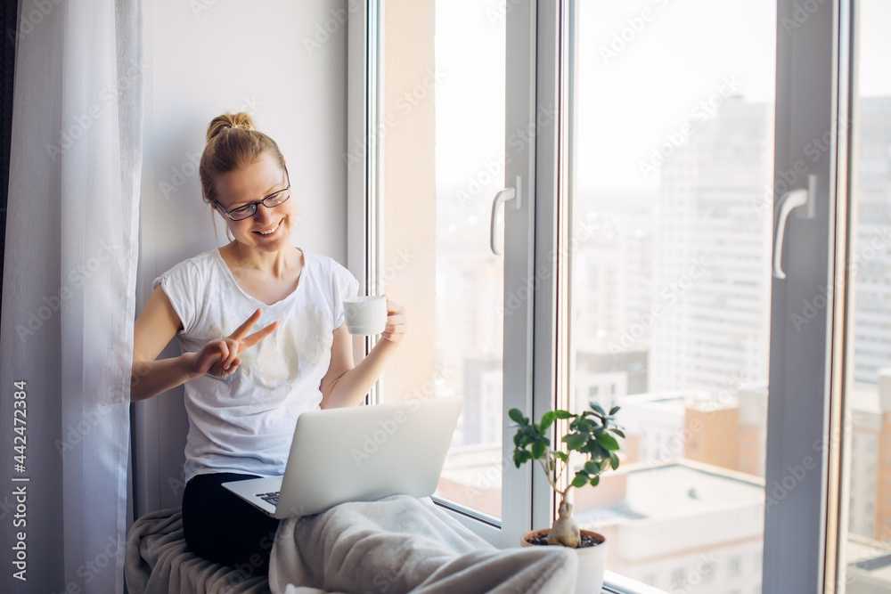 Happy smiling woman wearing glasses sitting on windowsill at city apartment, resting, looking at webcam on laptop, talking on video chat with her friends or family. Happy smiling woman wearing glasses sitting on windowsill at city apartment, resting, looking at webcam on laptop, talking on video chat with her friends or family.