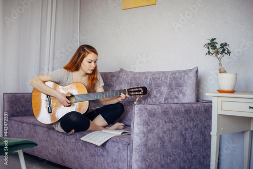 30 woman playing the guitar, sitting on the sofa in home interior. Lovely young girl learning to play the guitar with music sheets. Lifestyle, hobby, leisure, talent development concept.