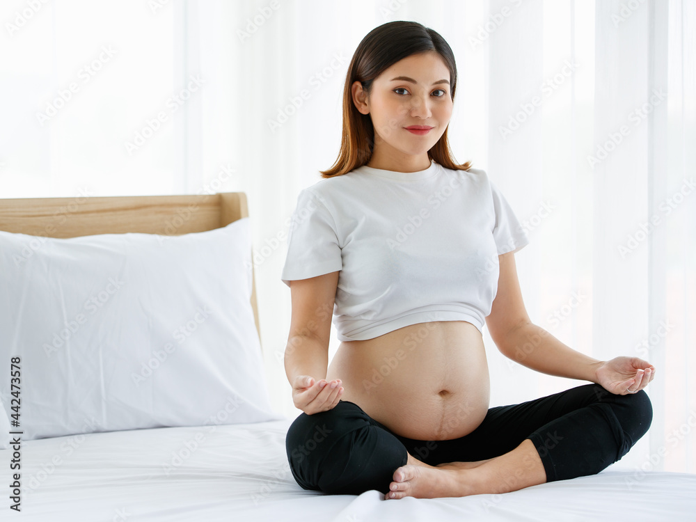 young pretty Asian pregnant woman sitting on a bed doing exercise delightfully by Yoga. She is smiling and looking at a camera. Healthy happy mother concept young pretty Asian pregnant woman sitting on a bed doing exercise delightfully by Yoga. She is smiling and looking at a camera. Healthy happy mother concept
