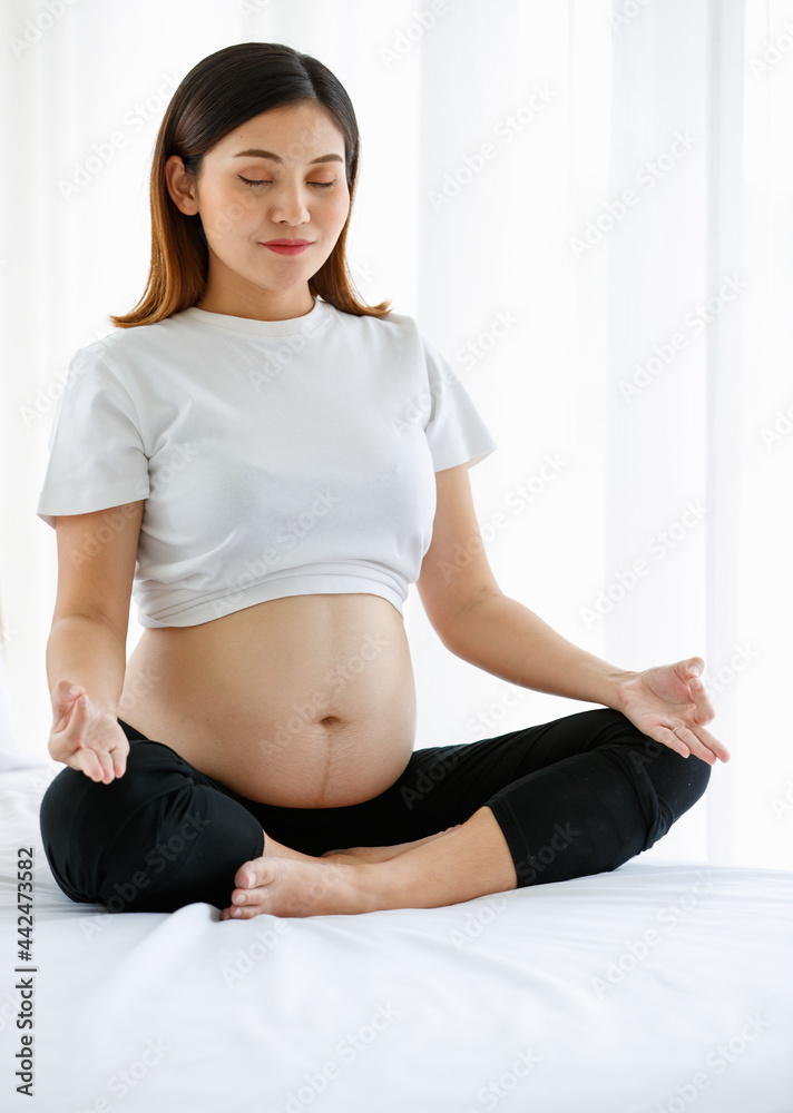 young pretty Asian pregnant woman sitting on a bed doing exercise delightfully by Yoga. She is smiling and looking at a camera. Healthy happy mother concept young pretty Asian pregnant woman sitting on a bed doing exercise delightfully by Yoga. She is smiling and looking at a camera. Healthy happy mother concept